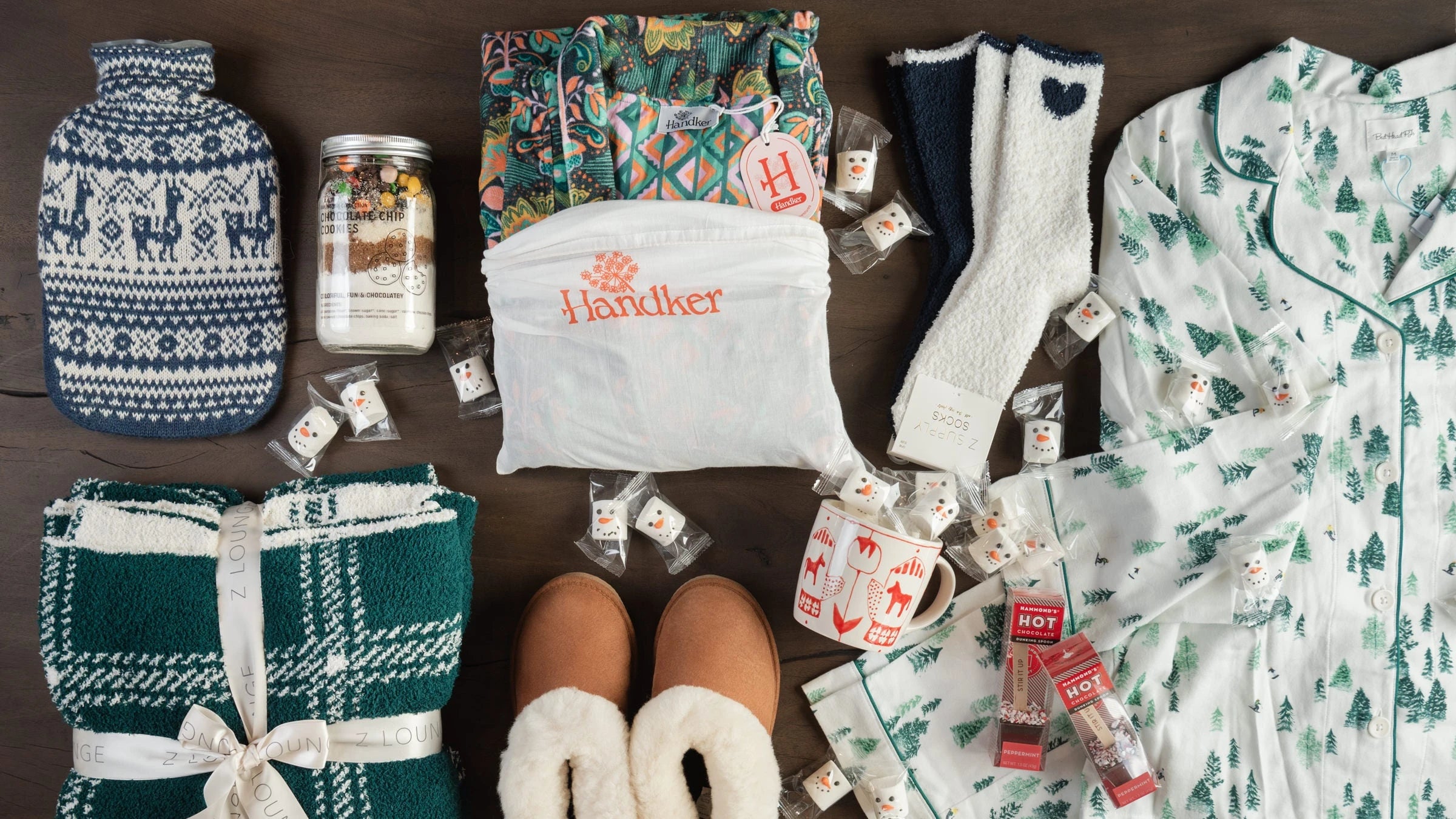 Cozy gifts including knitted hot water bottle cover, green plaid blanket, shearling slippers, cozy plush socks, and winter tree pajamas on a dark wood table at Addison West 