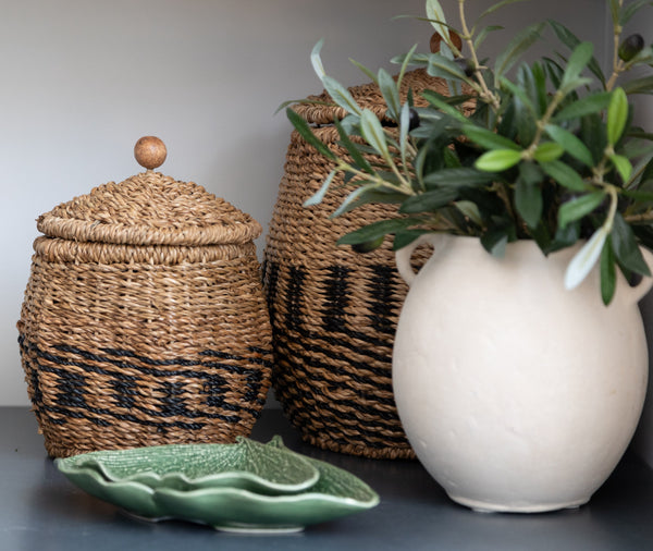 Pair of lidded woven baskets with lids on a shelf with a white vase and green small plates