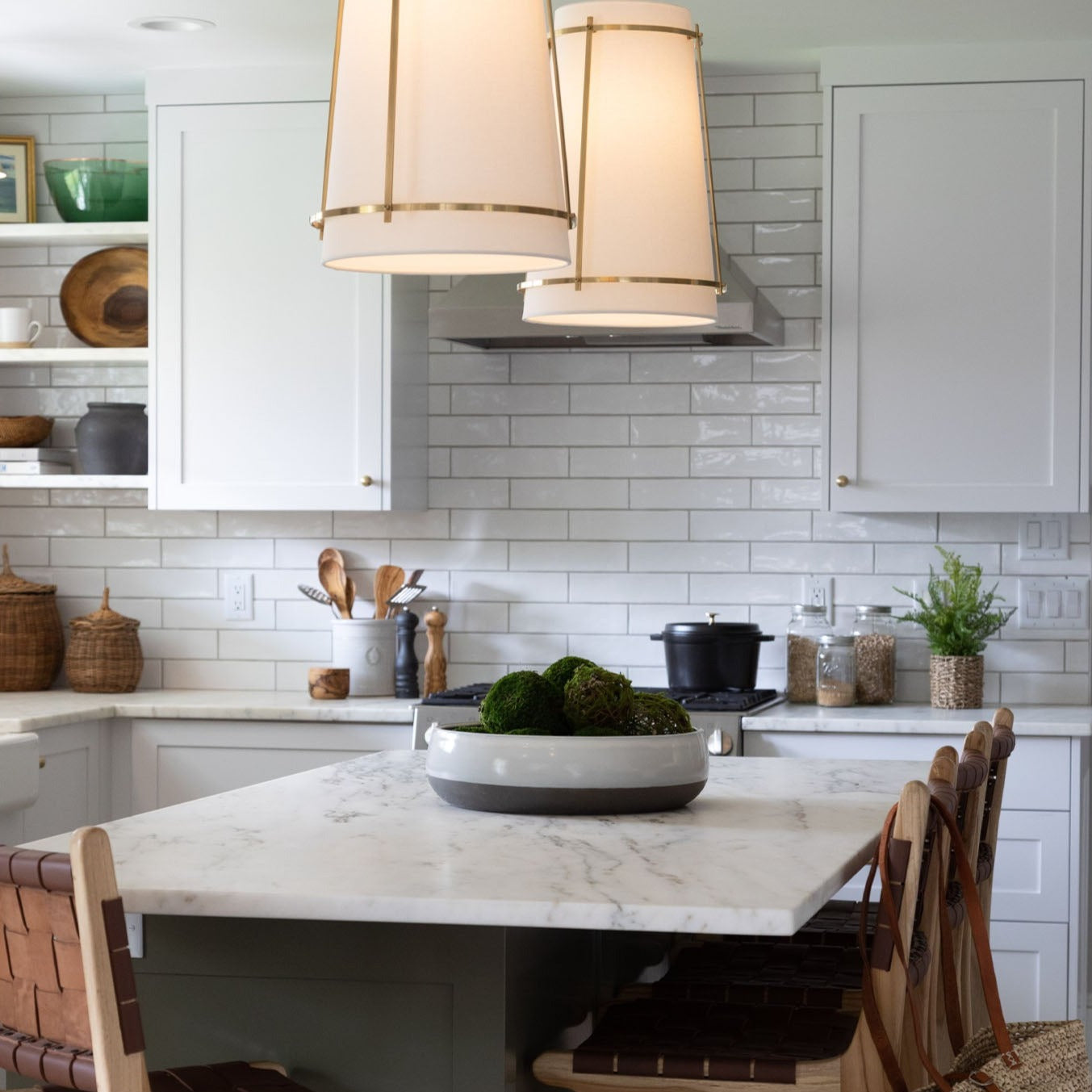 White kitchen with middle island and white pendants with large white bowl in the center filled with moss balls on a marble counter 