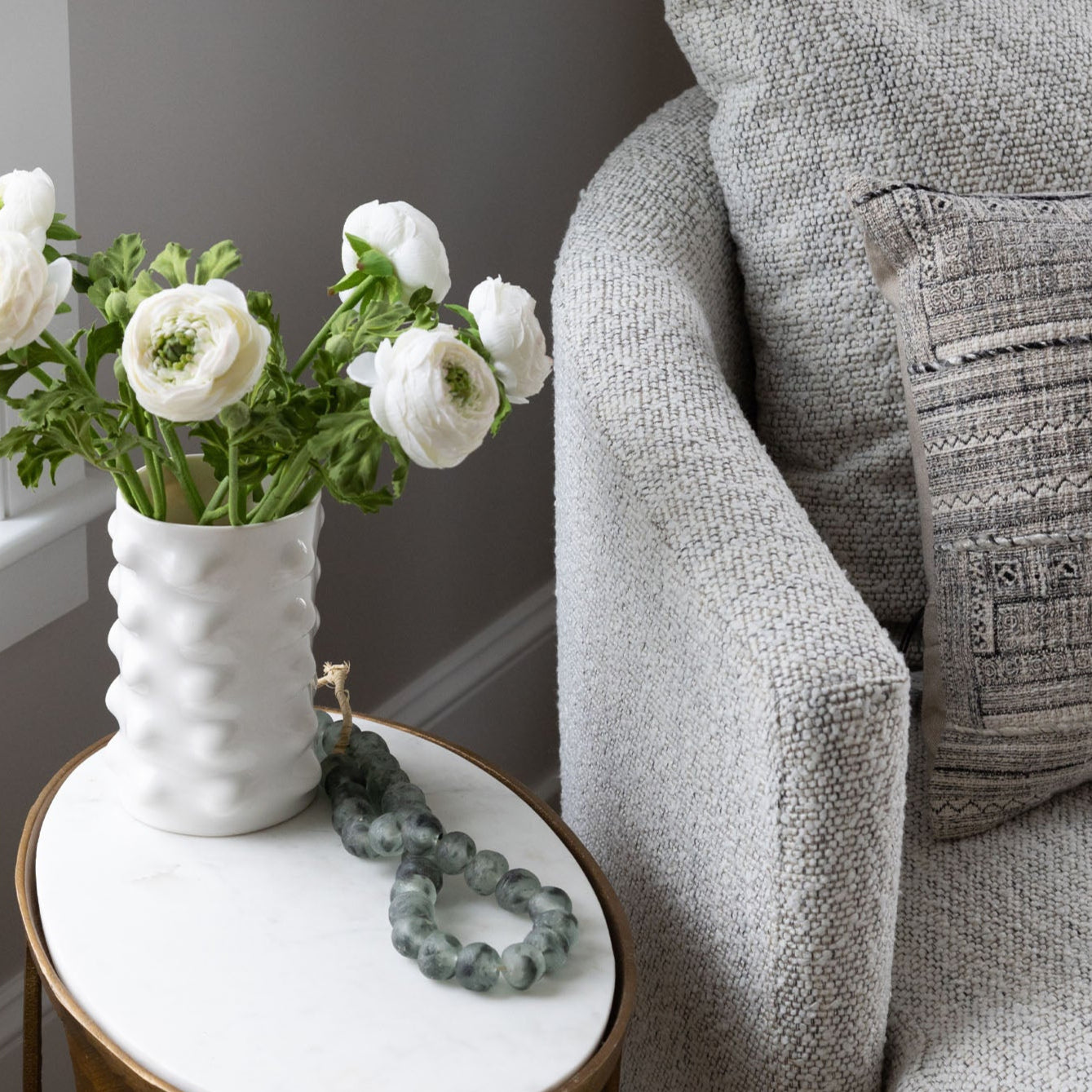 Vase of silk ranunculus flowers on a marble table beside an upholstered chair