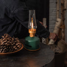 Light and Sound LED Lantern in Basil on a dark wood end table with a person stacking fire wood in the background