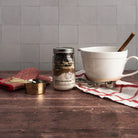 Chocolate Chip Rainbow Cookies Mix, ceramic mixing bowl with spout and handle, brass measuring cups, red oven mitts, and red and white kitchen towel on a wood table with a white tile background at Addison West