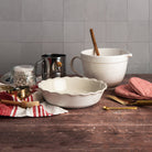 Emile Henry 9" Pie Dish in Sugar with ceramic mixing bowl with spout and handle, flour sifter, ceramic pie weights in a mason jar, red and white kitchen towel, pastry brush, brass spoon, gold pie server on a wood table with a white tile background at Addison West