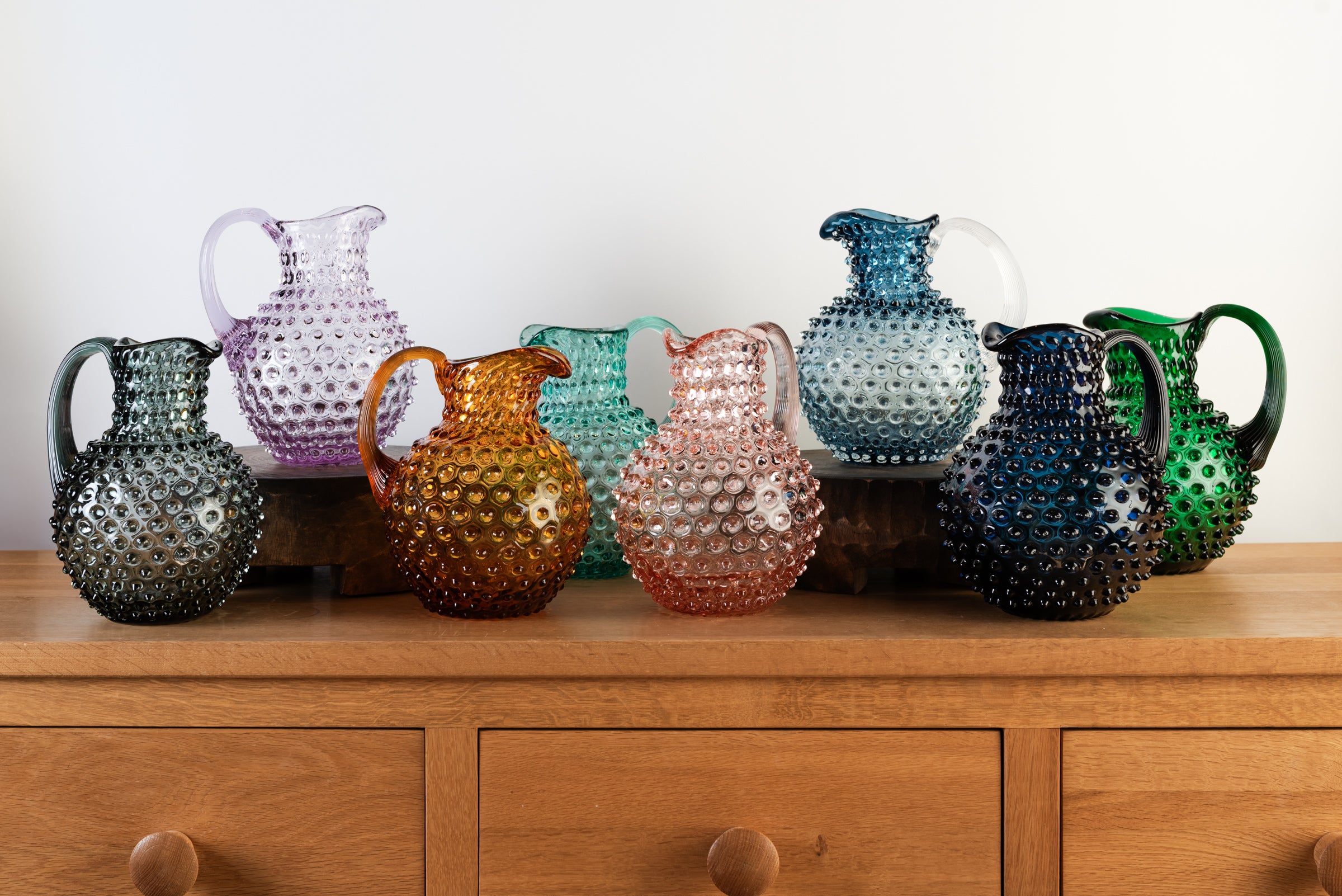 Collection of glass hobnail pitchers on a wood table in front of a white wall at Addison West