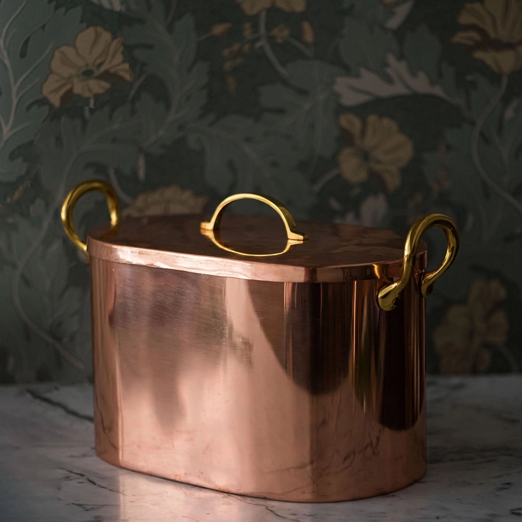 Copper bread box with three brass handles on a marble counter top with floral wallpaper in the background