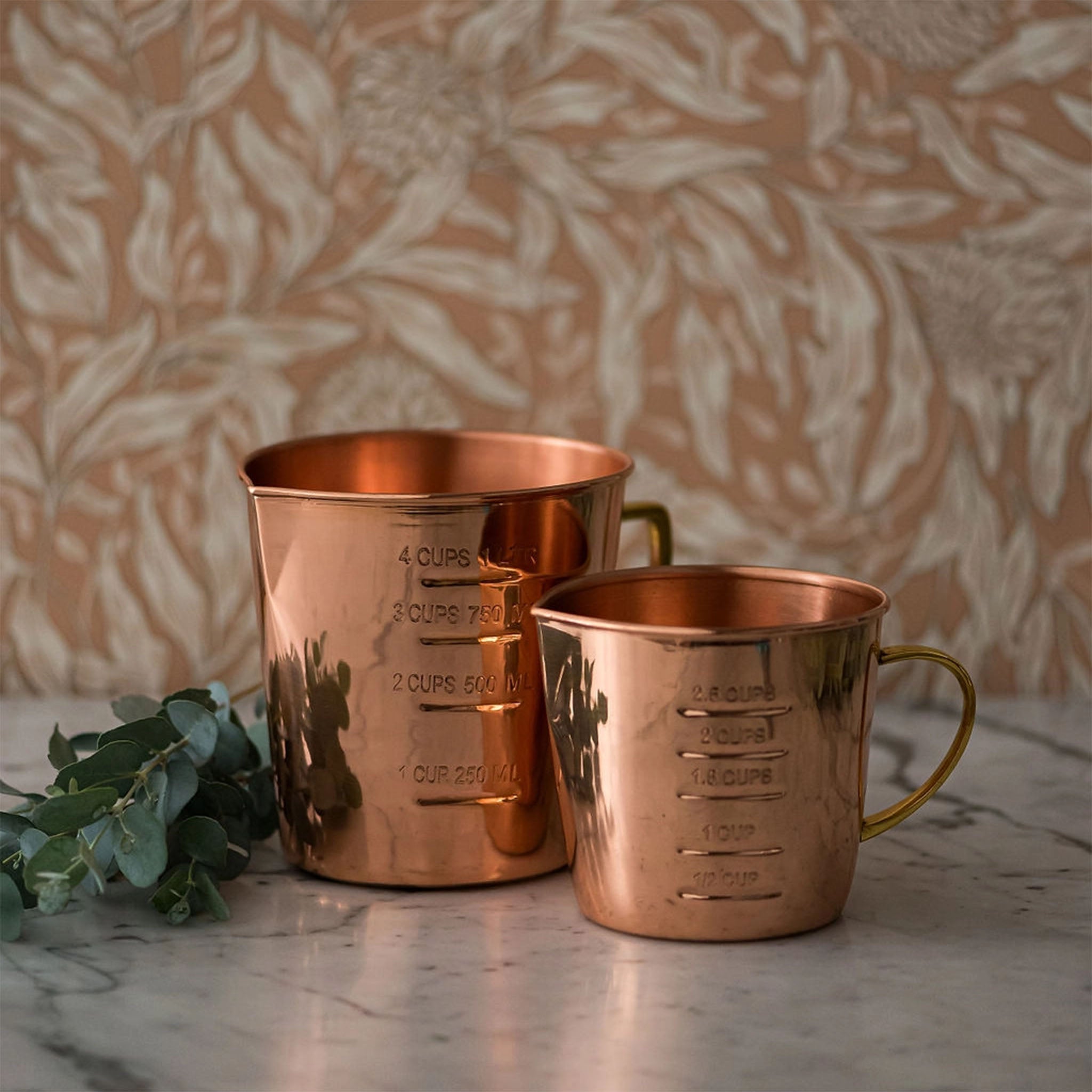 Two copper measuring cups on a marble counter top with floral wallpaper in the background