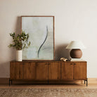 Four Hands Eaton Media Cabinet in Amber Oak Resin in a living space with beige walls and a painting propped on top