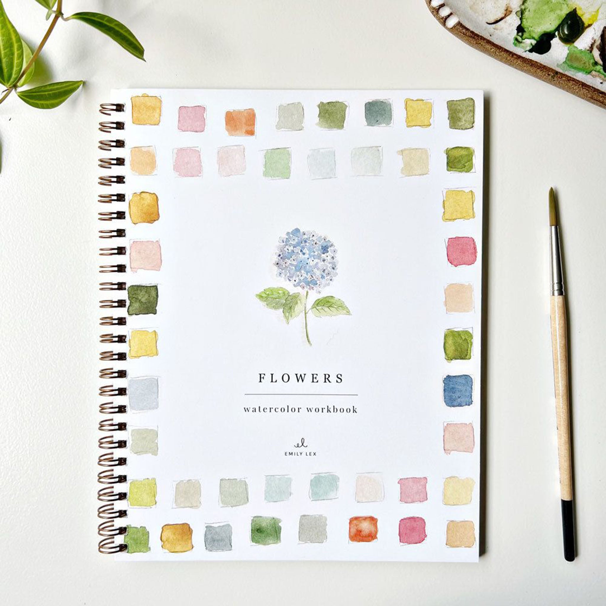 Emily lex studio Flowers Watercolor Workbook on a white surface with a paint brush and greenery