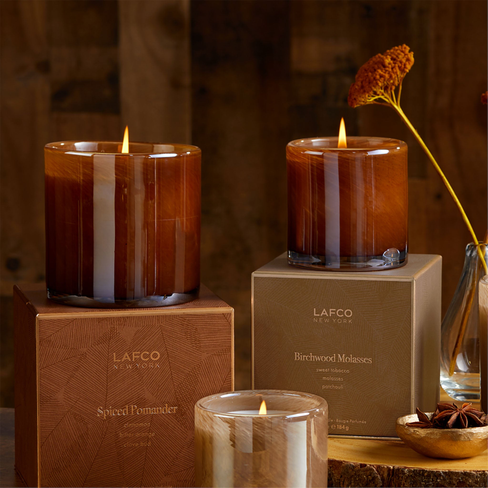 Brown candle and boxes of Lafco's spiced pomander scent on a wood table in front of a wood wall