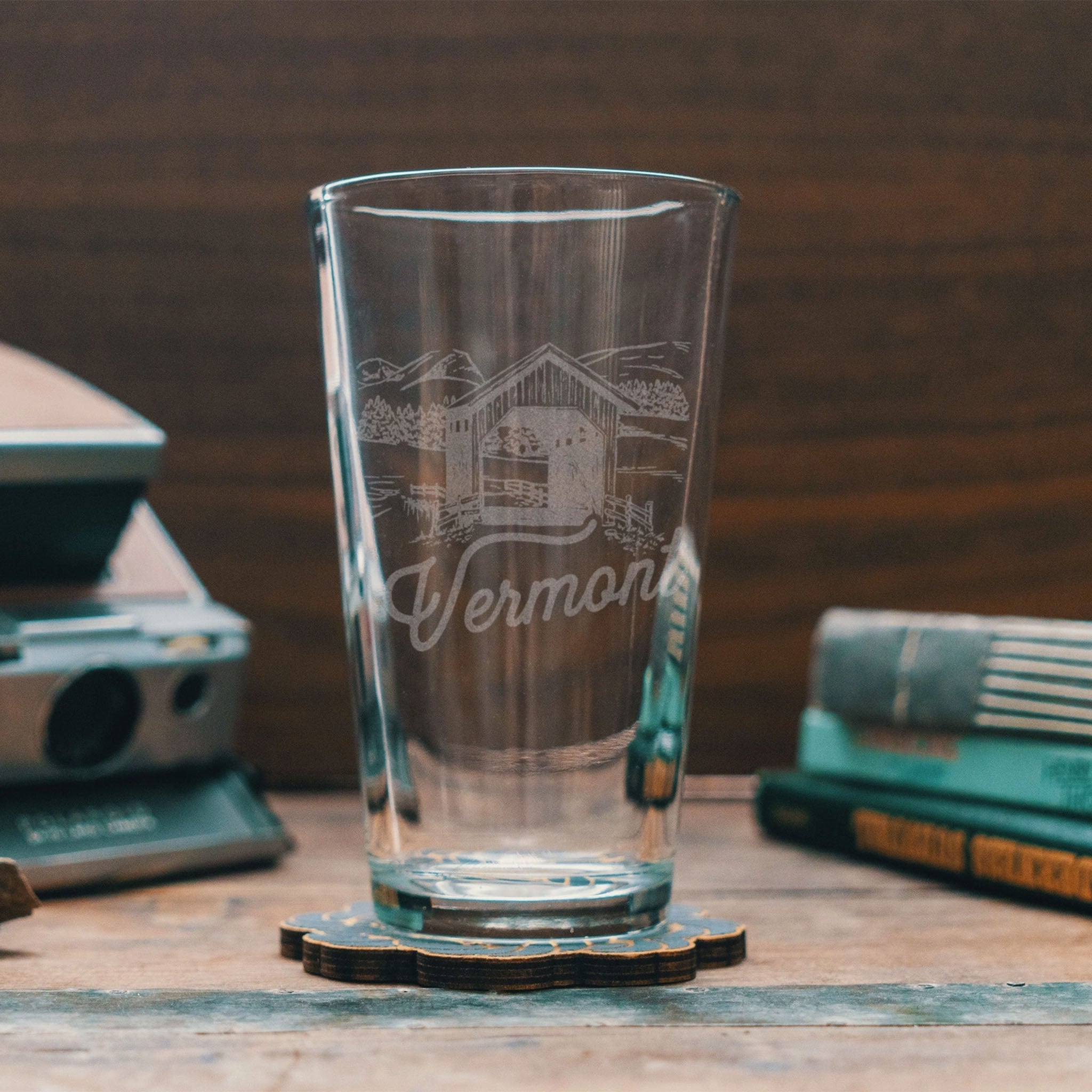 Vermont pint glass with etched image of a covered bridge on a wood surface next to green books.