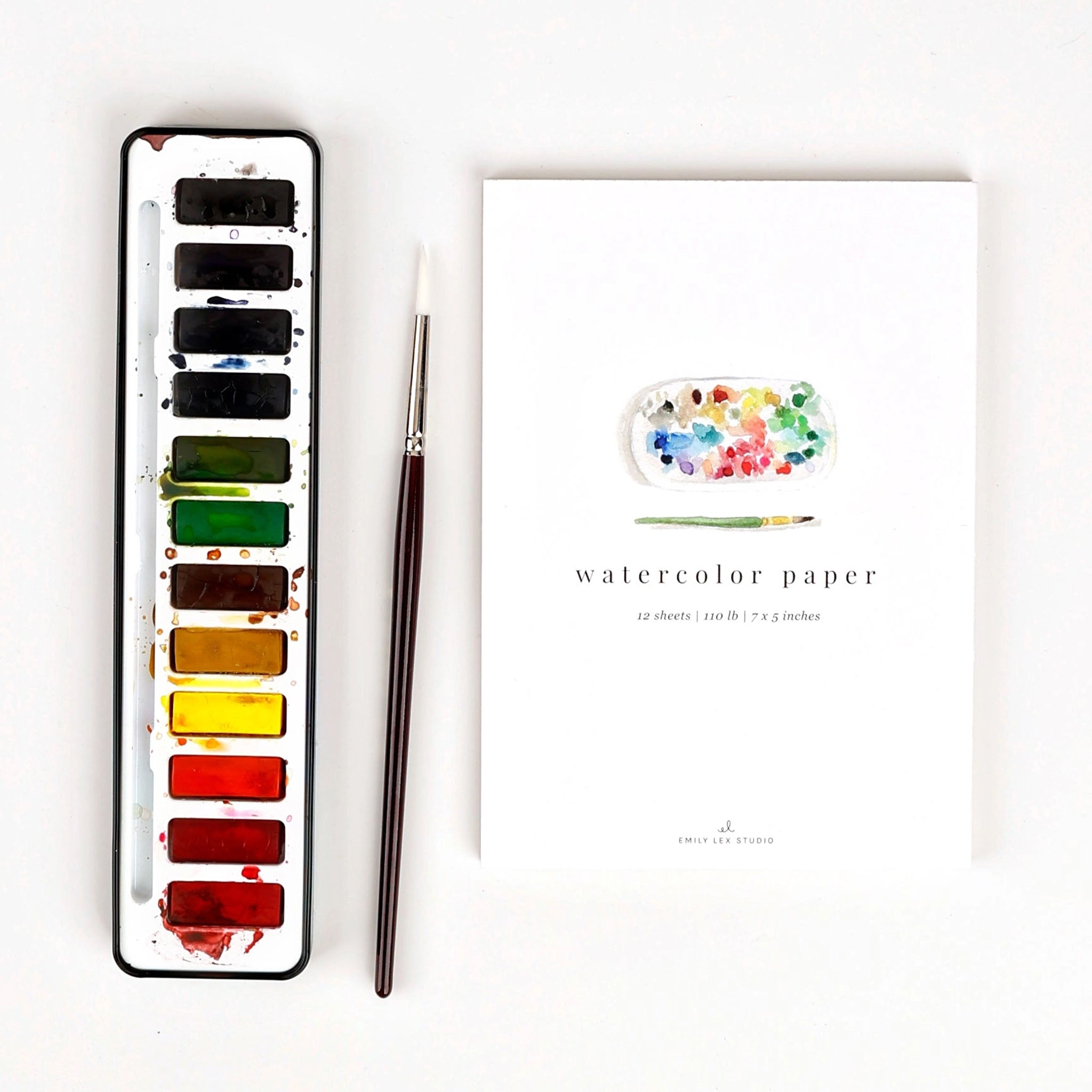 Set of water color paints with brush next to watercolor paper on a white background