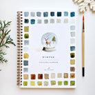 Emily Lex Winter Watercolor Workbook on a white surface next to a brush and greenery