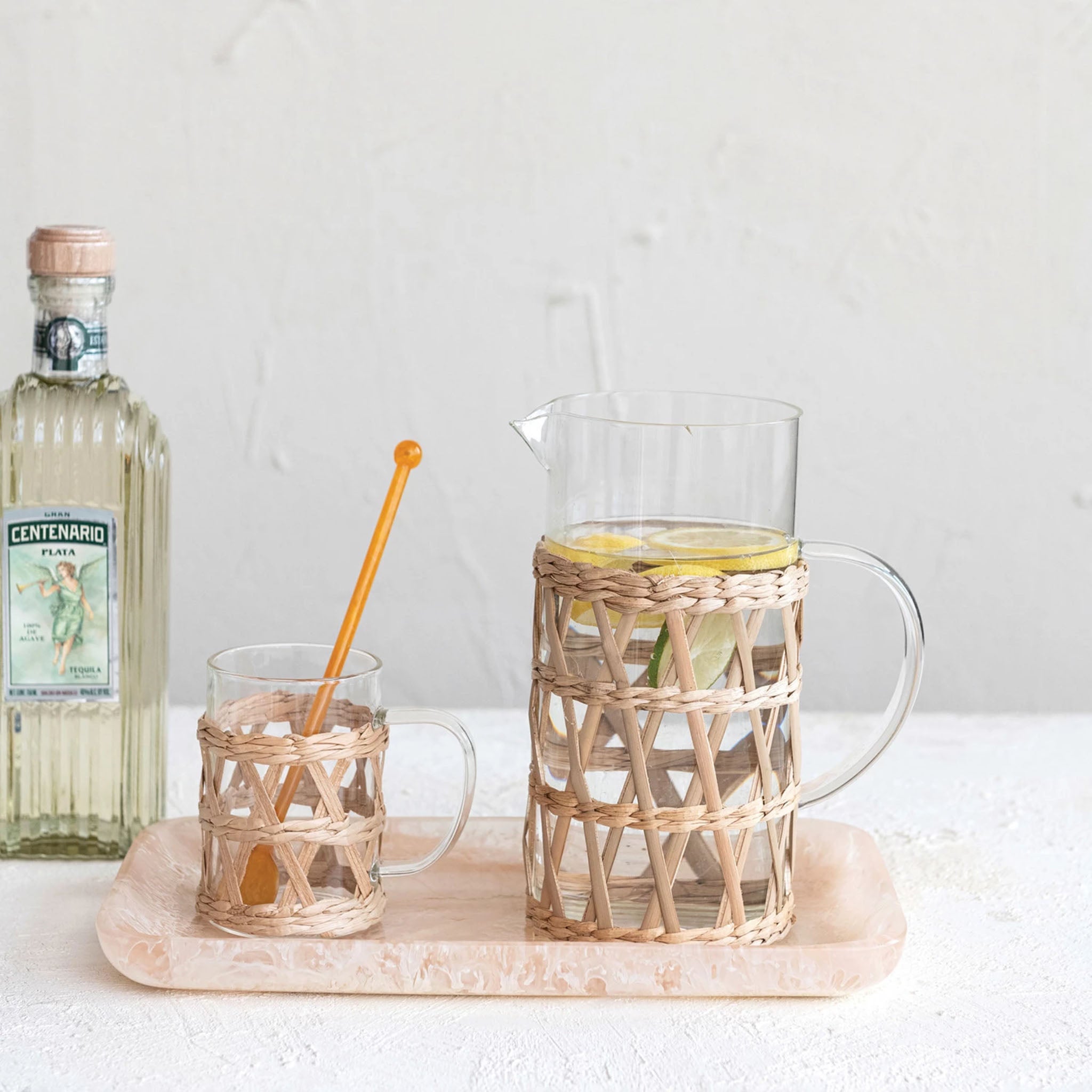 Seagrass Wrapped Glass Pitcher filled with liquid and lemons on a counter with glass liqour bottle 