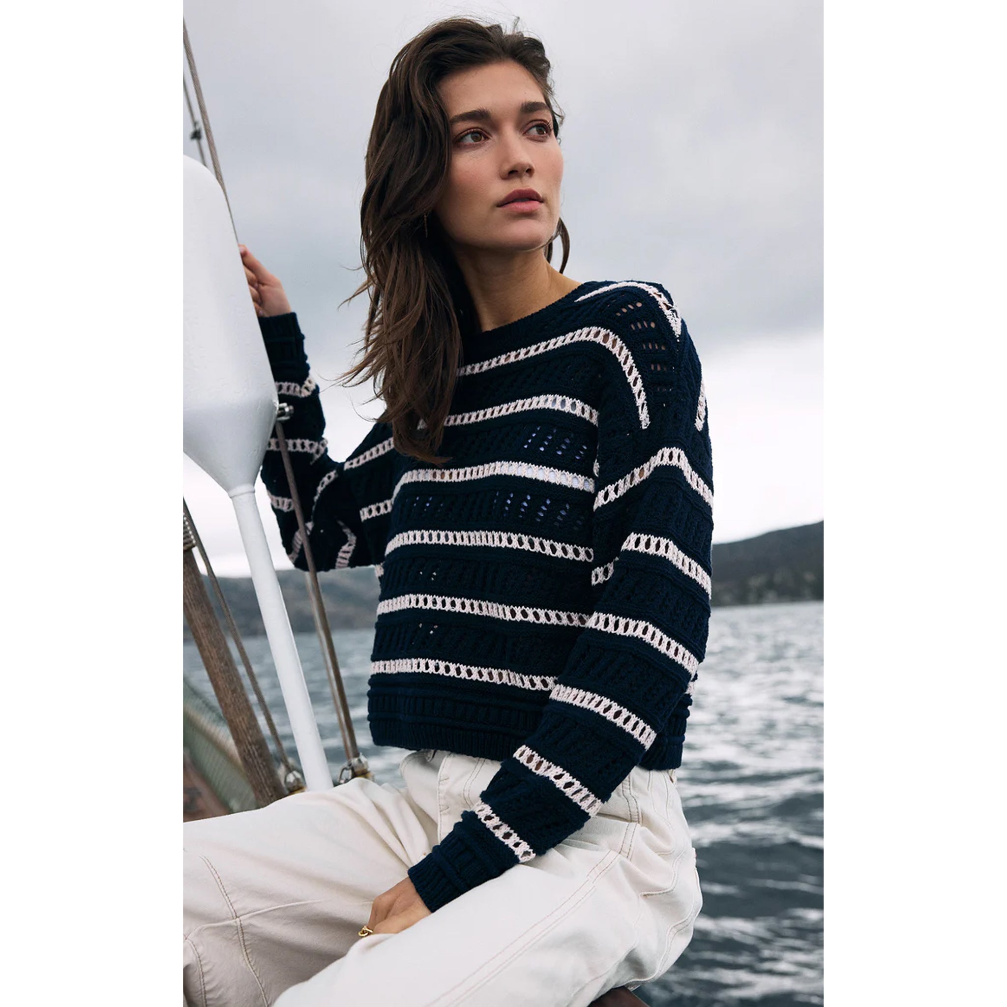 Model wearing Z Supply Estero Stripe Sweater Eclipse with white jeans on a boat on a lake or ocean