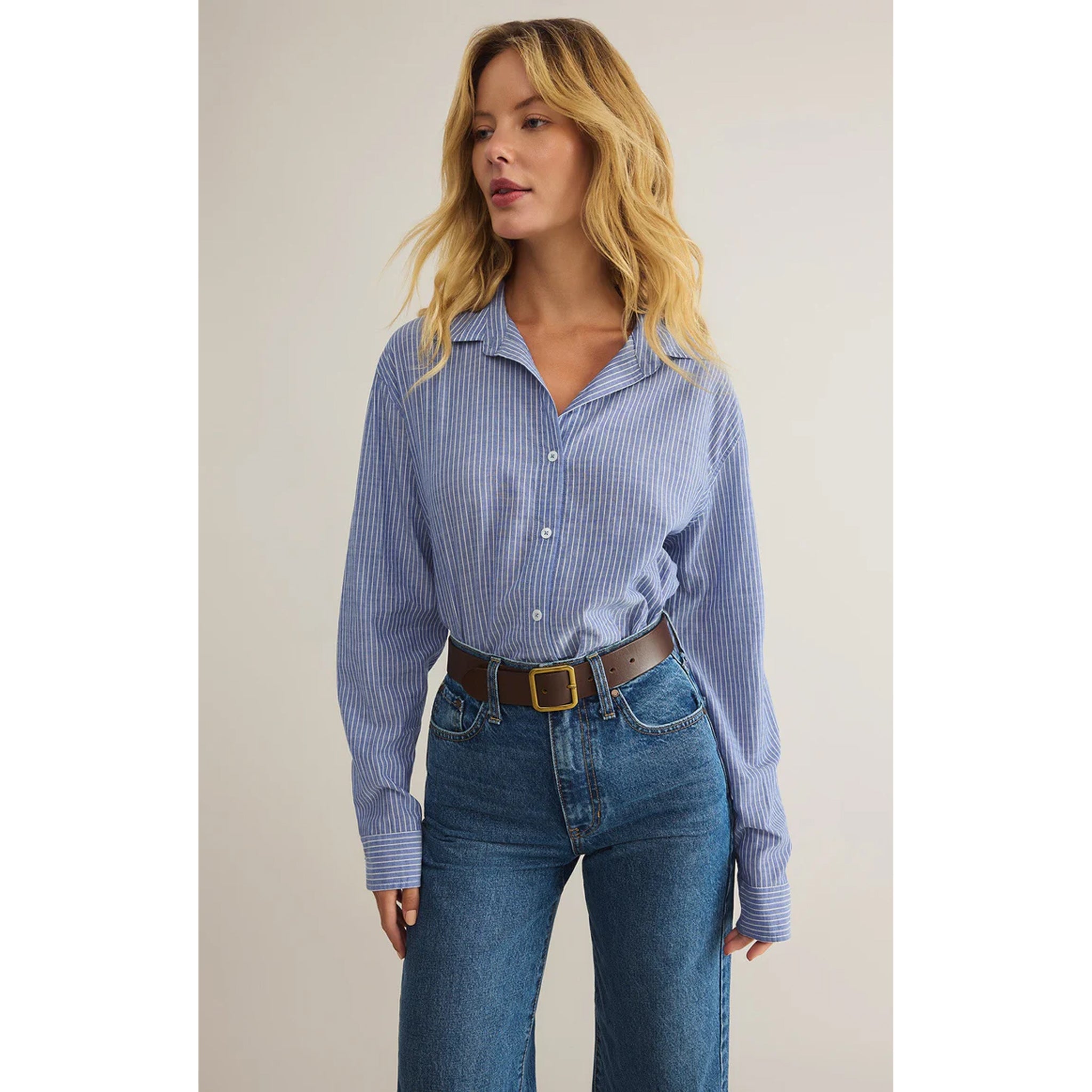 Model wearing Z Supply Seaport Striped Shirt in Sky Blue tucked in with blue jeans and a brown belt in front of a neutral backdrop