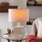 Surya brand Bentley table lamp with glass base and beige linen shade on a side table next to a chair and white brick fireplace