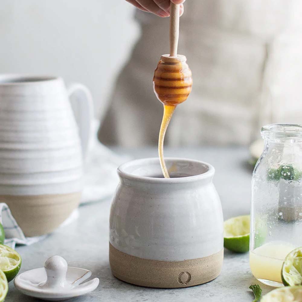 Farmhouse Pottery Beehive Honey Pot on a table with limes and a person holding a honey dipper dripping honey