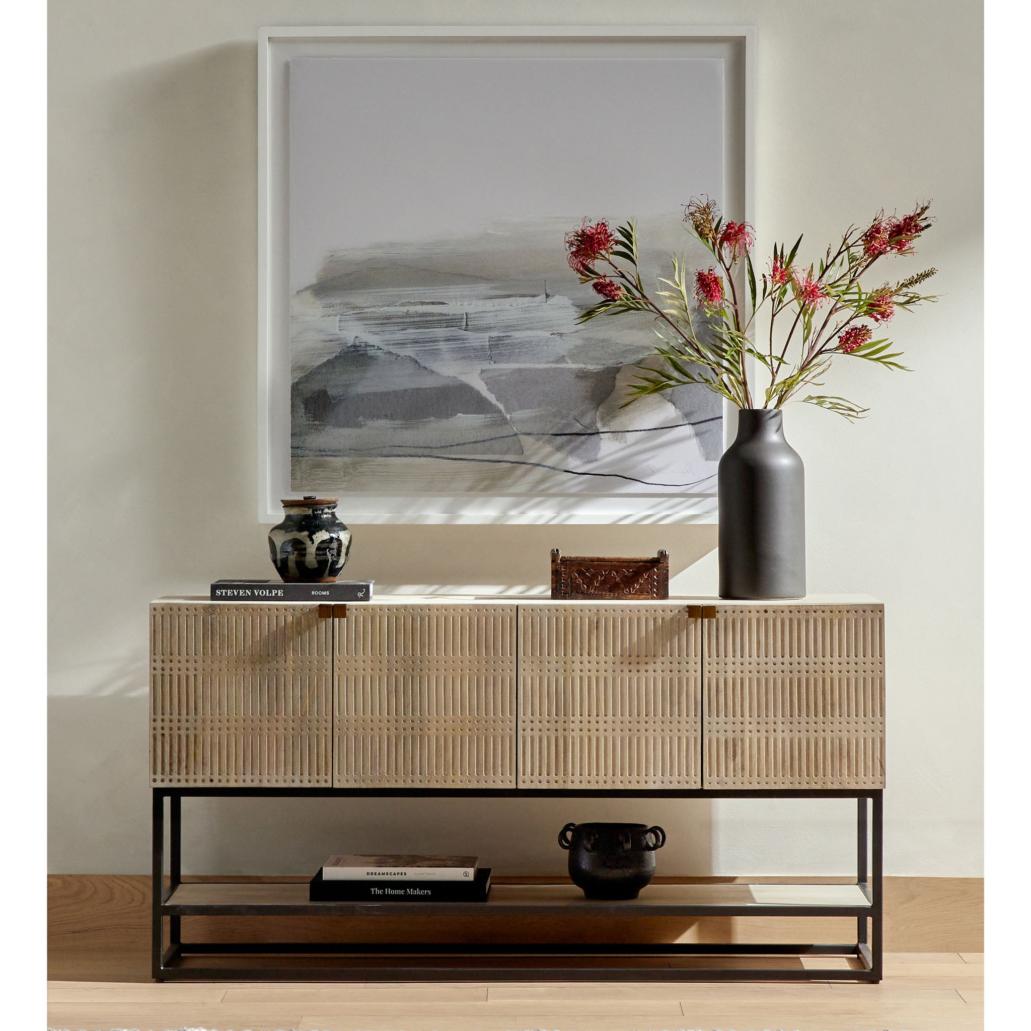 Living space with large square artwork and carved front console with iron base and shelf in front of a cream wall 