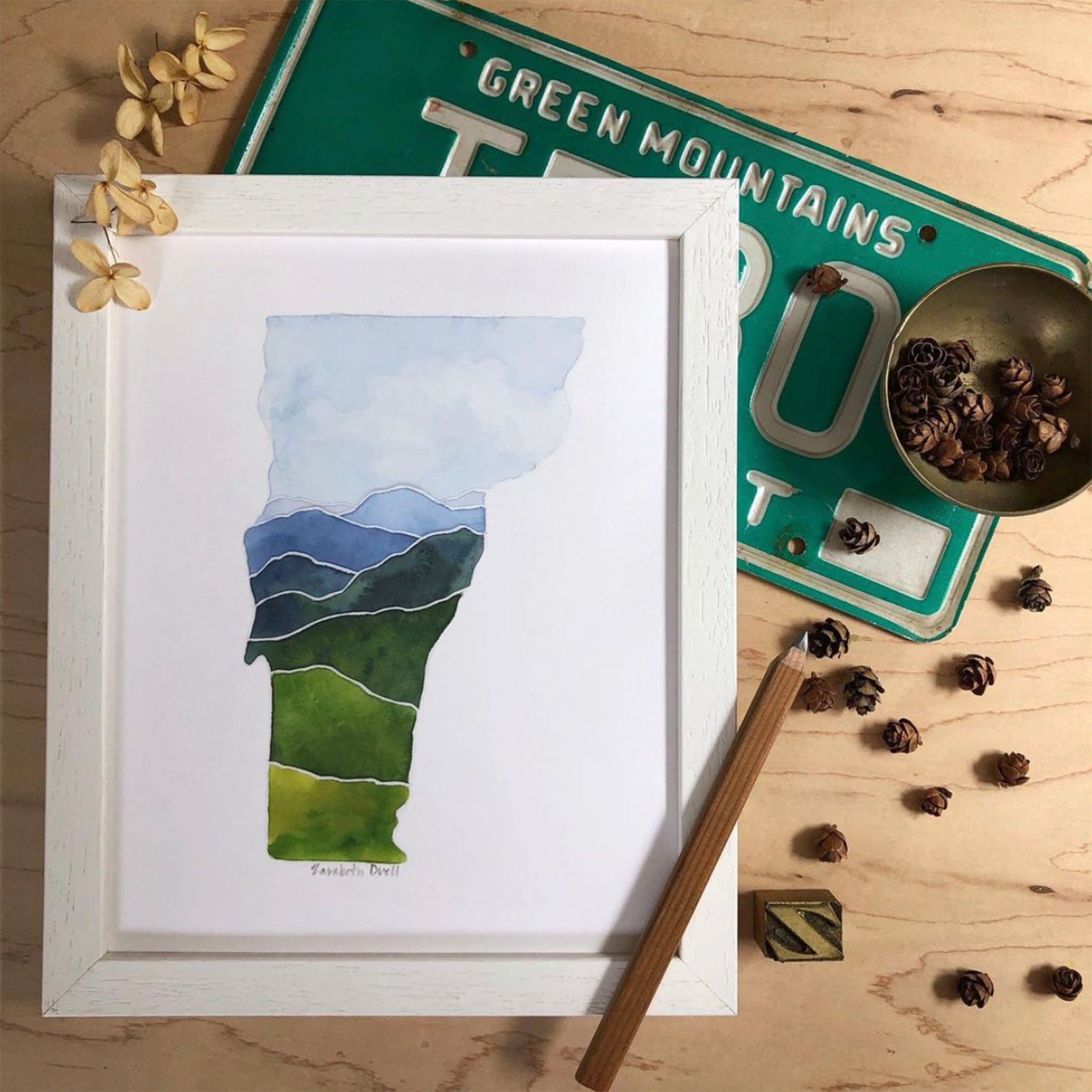 Print of Vermont state shape in green and blue watercolors in a white frame on a wood table with pinecones by Zarabeth Duell 