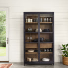 Four Hands furniture brand Millie black cabinet with two glass doors and oak interior on a concrete floor filled with plates and glasses against a white wall 