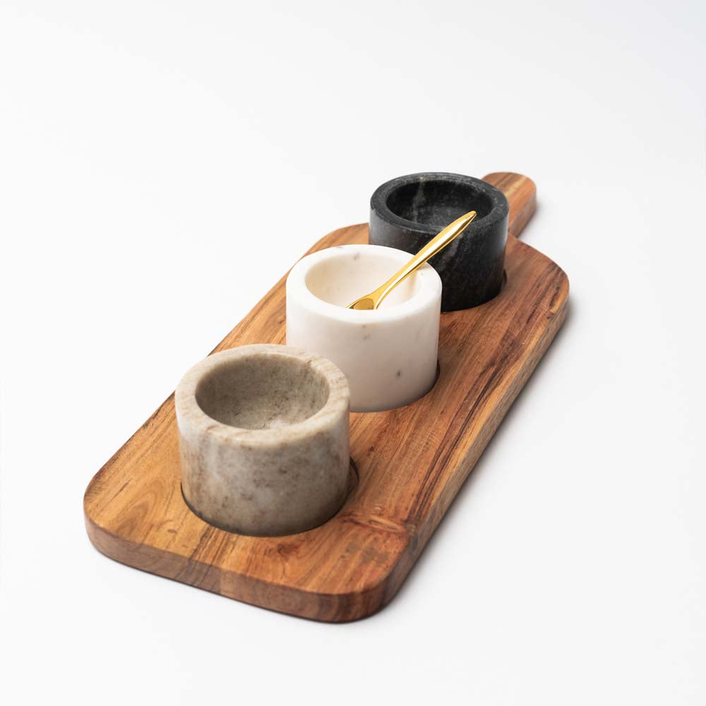 Wood serving board with three marble pinch pots and a brass spoon on a white background