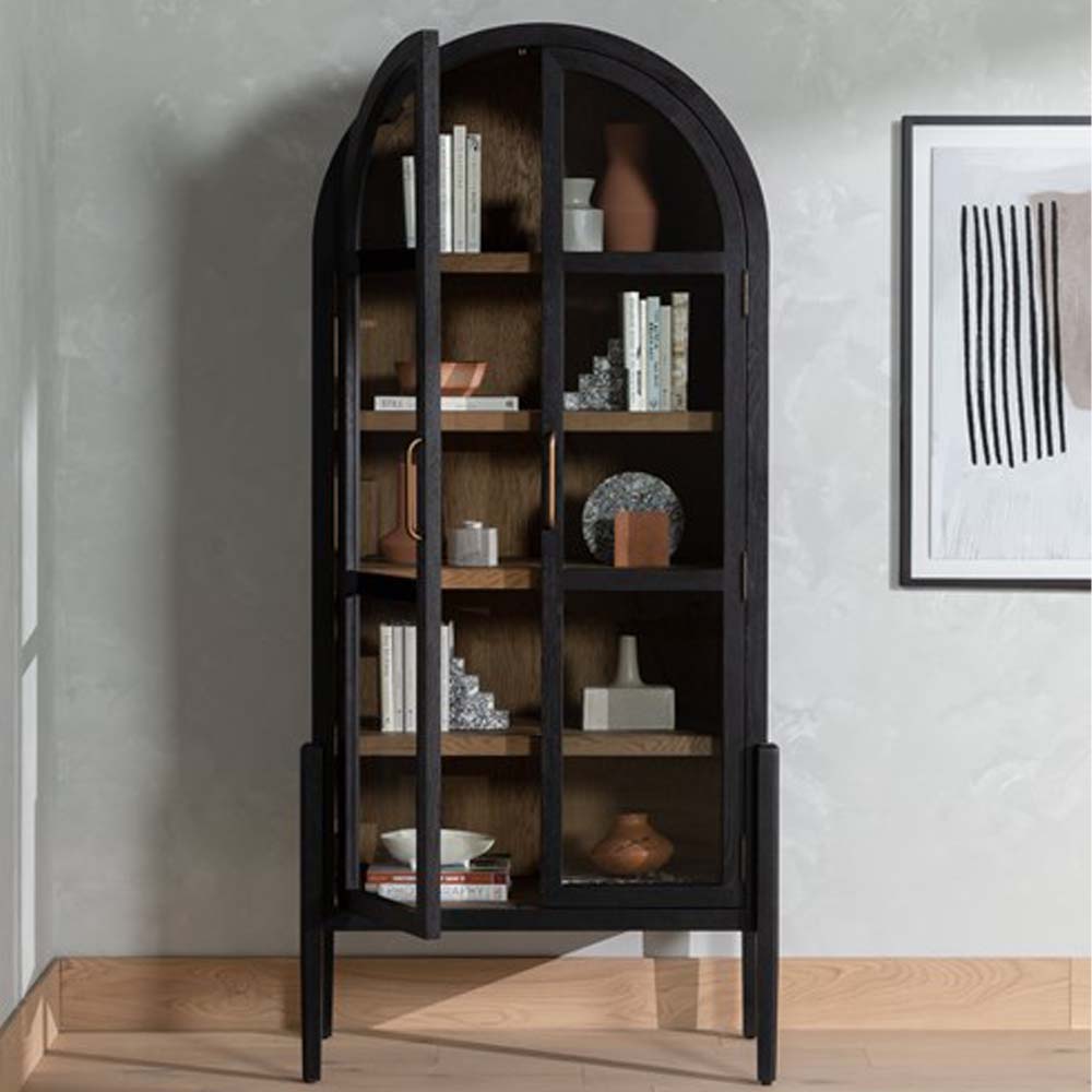 Four hands furniture brand Tolle black cabinet with arched top and wood stained interior and two glass doors with decorative items in a room with wood floor and white wall